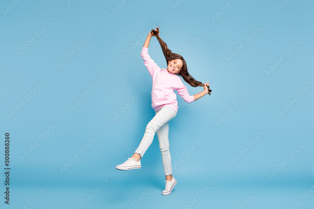 Full length body size turned photo of cheerful positive funny child smiling toothily jumping in happiness pink hoodie pulling her hair isolated blue pastel color background