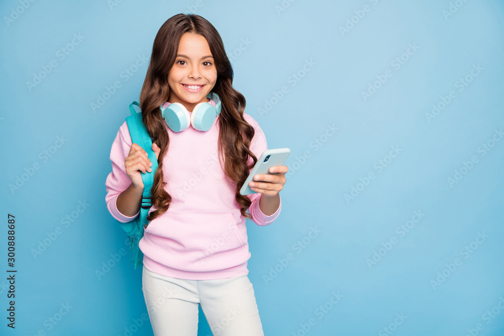 Photo of pretty small school lady hold telephone turning on popular youth song wireless earflaps wear rucksack casual hoodie hood pink pullover isolated blue color background - obrazy, fototapety, plakaty 