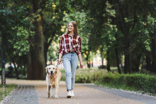 full length view of beautiful girl in casual clothes waking in park with golden retriever