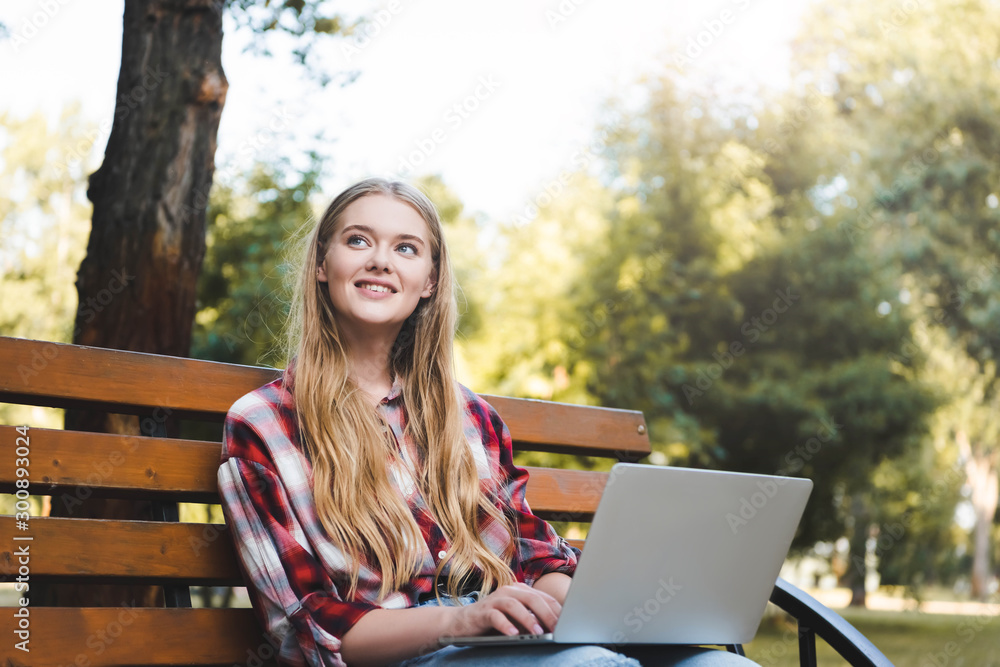 beautiful girl in casual clothes sitting on wooden bench in park, using laptop and looking away