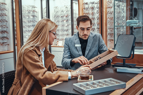 Woman is choosing eyeglasses while male optician sitting near with another eyeglasses. photo