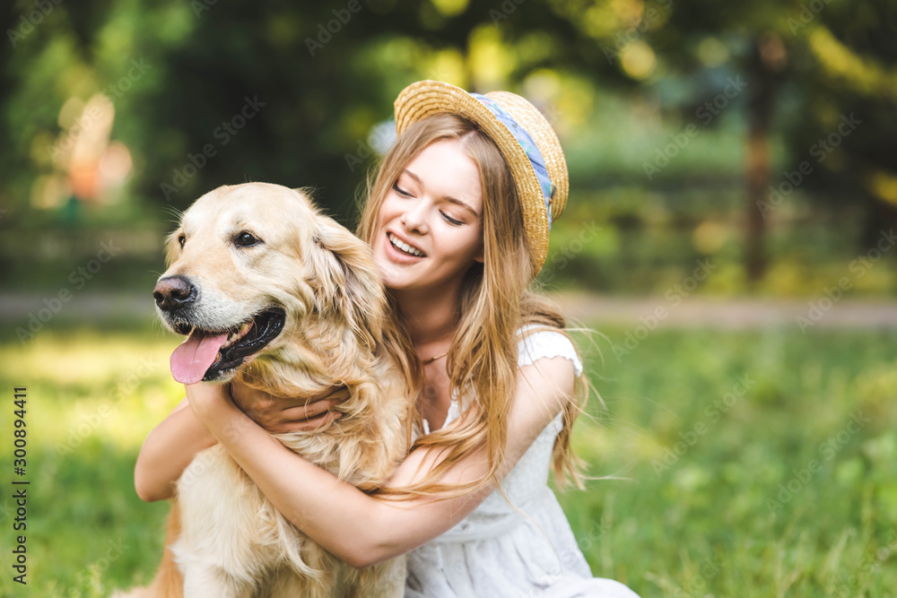 selective focus of beautiful girl in white dress and straw hat hugging golden retriever and smiling while sitting on meadow selective focus of beautiful girl in white dress and straw hat hugging golden retriever and smiling while sitting on meadow