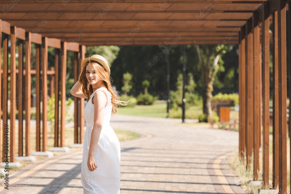 beautiful girl in white dress touching straw hat while walking near wooden construction and looking at camera