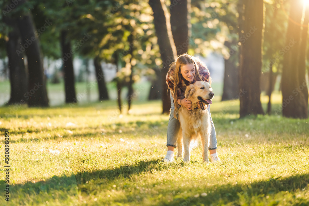 full length view of beautiful girl in casual clothes hugging golden retriever while standing on meadow in sunlight