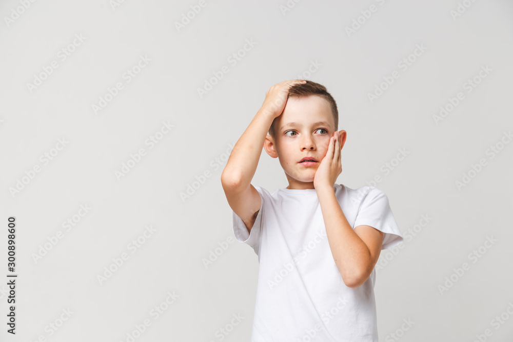 Scared boy in white t-shirt with both hands on head on gray background Scared boy in white t-shirt with both hands on head on gray background