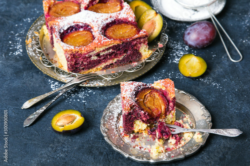 Tasty plum cake with pieces of fruit and powdered sugar on a dark background