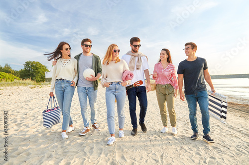 friendship, leisure and people concept - group of happy friends with ball, guitar, bag and picnic basket with blanket walking along beach in summer