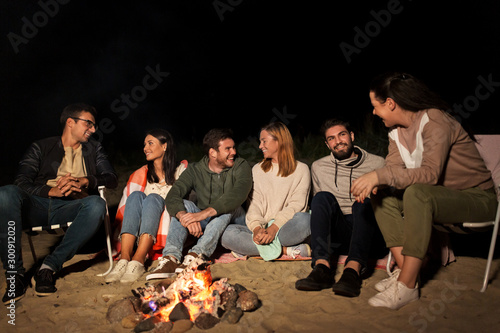 leisure and people concept - group of smiling friends sitting at camp fire on beach at night