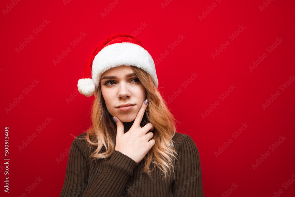 Portrait of pensive girl on red background, looks away and thinks wearing warm sweater and hat. Lady in santa hat looks away with a thoughtful look, copy space. Isolated. Portrait of pensive girl on red background, looks away and thinks wearing warm sweater and hat. Lady in santa hat looks away with a thoughtful look, copy space. Isolated.