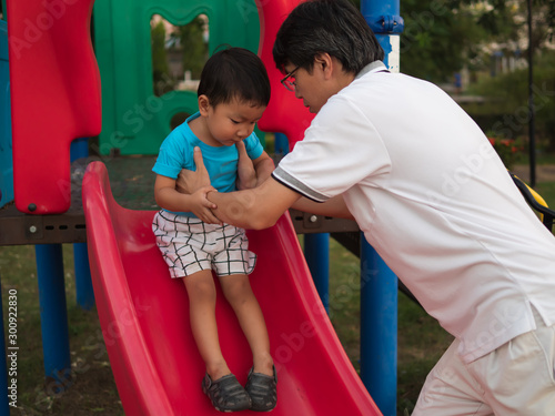 Asian child boy playing slider in playground together with dad in happy family time. Father support son with love and care.