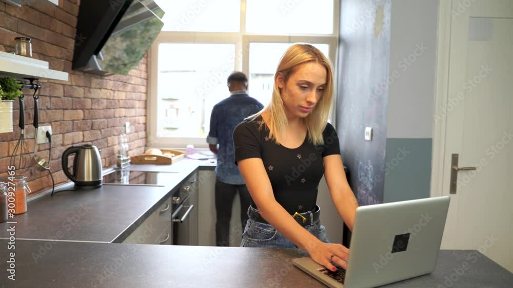 Young woman working at the notebook while boyfriend washes the dishes. Young woman working at the notebook while boyfriend washes the dishes.