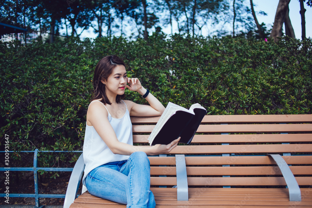 Naklejka premium Young Asian concentrated lady reading book and sitting on bench against hedge