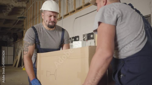 Two strong muscular Caucasian workers taking up heavy cardboard box and carrying it at the construction site. Adult male builders in protective helmets and uniform working indoors. Hard work. photo