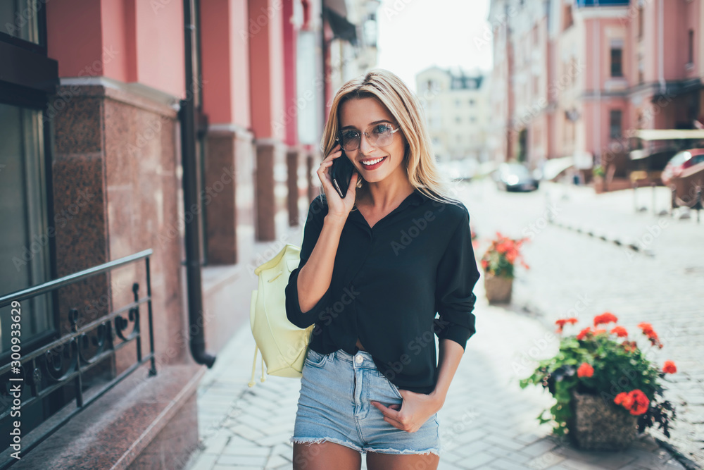Fototapeta premium Half length portrait of positive carefree female tourist in trendy sunglasses smiling during smartphone conversation with contact service, positive woman talking via cellphone standing outdoors
