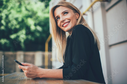 Concept of technology and generation, attractive female with candid smile on face looking away while resting outdoors, positive blonde hipster girl holding cellular phone for online communication
