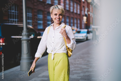Half length portrait of cheerful female traveller with backpack and smartphone device in hand looking at camera while standing on modern urban street, happy smiling hipster girl resting outdoors