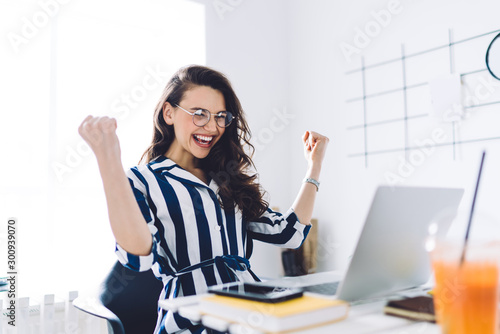 Happy young woman sitting at table with hands up in winner gesture