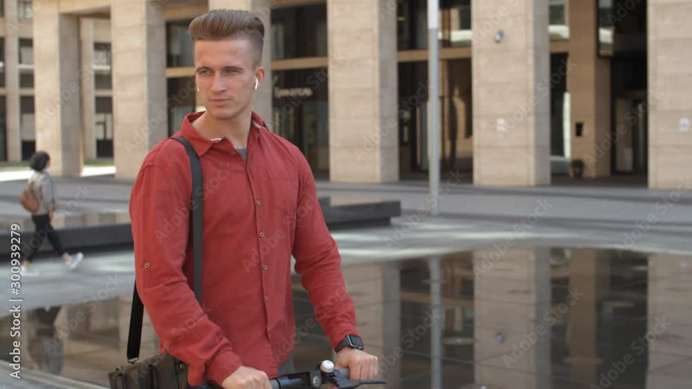 Tilt down of young good-looking Caucasian man wearing trendy clothes and smart watch riding electric kick scooter in city while listening to music through wireless headphones Tilt down of young good-looking Caucasian man wearing trendy clothes and smart watch riding electric kick scooter in city while listening to music through wireless headphones