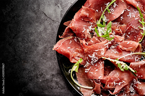 Tasty appetizing classic Italian bresaola with rocket salad and parmesan on black plate on black background. View form above. Top view. Horizontal
