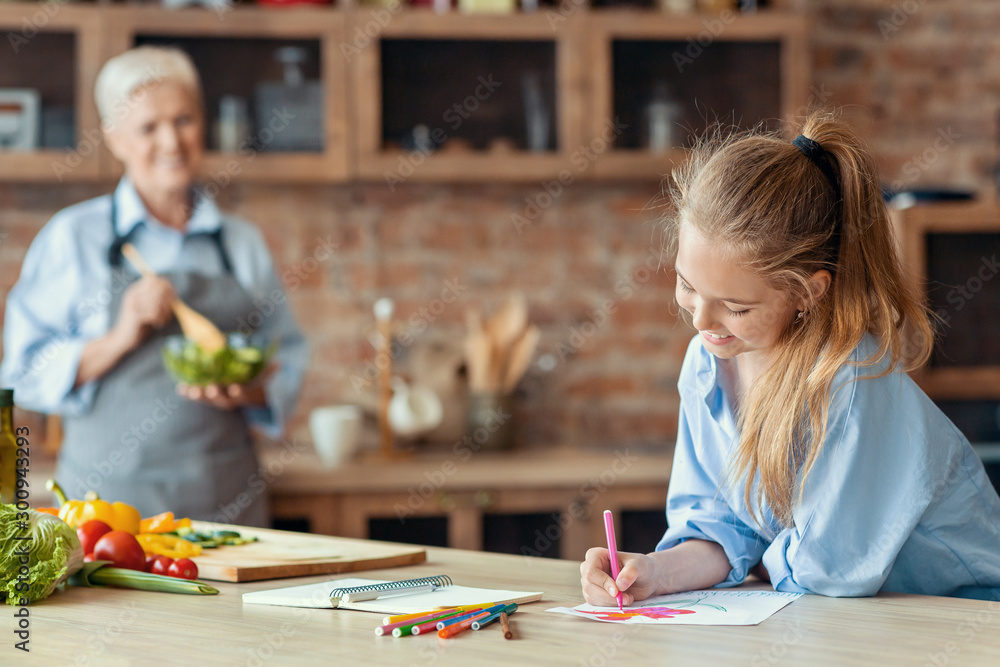 Fototapeta premium Little girl drawing at kitchen while helping granny