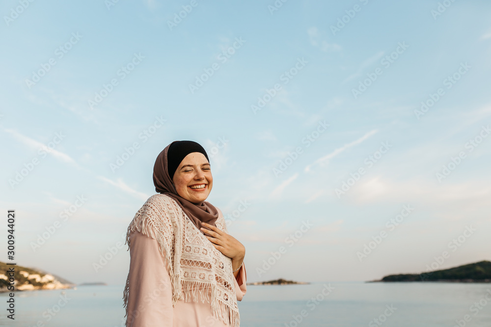 portrait of young European Muslim women with hijab sitting on the beach with hat in her hand. She is happy and relaxed. Sea is in the background. She is looking at the camera. portrait of young European Muslim women with hijab sitting on the beach with hat in her hand. She is happy and relaxed. Sea is in the background. She is looking at the camera.