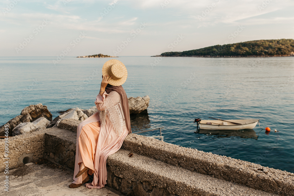 portrait of young European Muslim women with hijab sitting on the beach with hat in her hand. She is happy and relaxed. Sea is in the background. She is looking in the distance. portrait of young European Muslim women with hijab sitting on the beach with hat in her hand. She is happy and relaxed. Sea is in the background. She is looking in the distance.