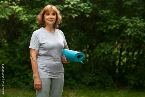 European woman 60 years old with fitness mat in summer park  copy space. Mature woman getting ready for outdoor sports. Active and healthy lifestyle.