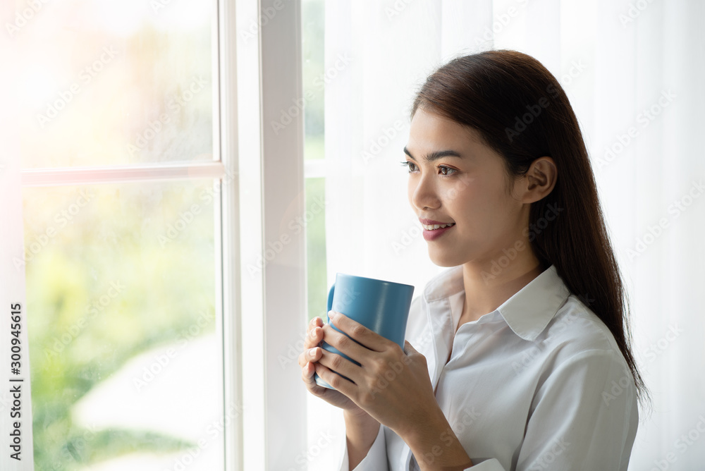 Asian woman drinking a cup of coffee and looking through the glass window after wake up in the morning Asian woman drinking a cup of coffee and looking through the glass window after wake up in the morning