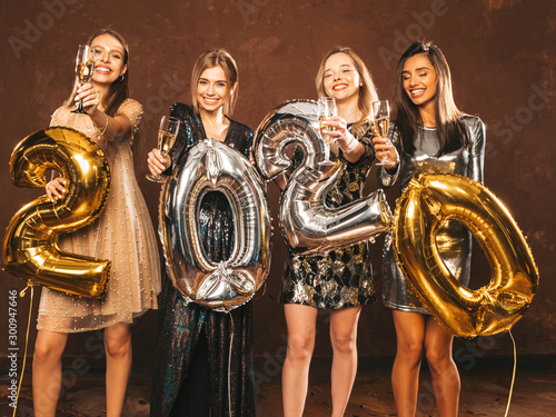Beautiful Women Celebrating New Year.Happy Gorgeous Girls In Stylish Sexy Party Dresses Holding Gold and Silver 2020 Balloons, Having Fun At New Year's Eve Party.Сarrying and drinking champagne flutes