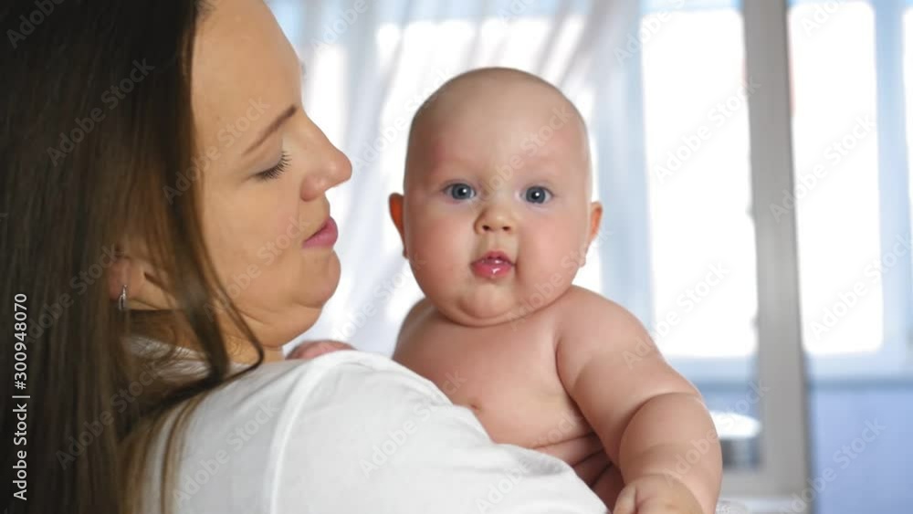 Mother holds a chubby smiling baby in her arms at home. The kid is looking at the camera. Close up shot. Mother holds a chubby smiling baby in her arms at home. The kid is looking at the camera. Close up shot.