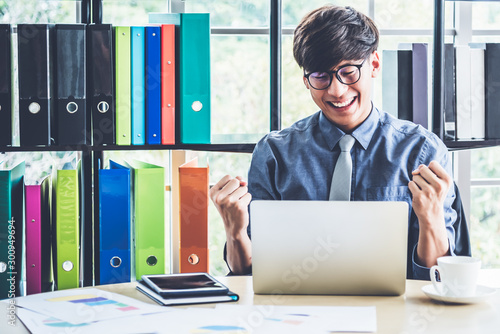 Asian business men Is smiling and happy when he looks at the earning Successful From a notebook computer on wooden table and with Business docoments placed on shelves background.