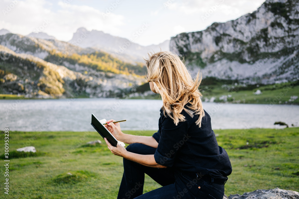 Naklejka premium A beautiful caucasian blonde woman writes and draws on a notepad in a mountain landscape with lake