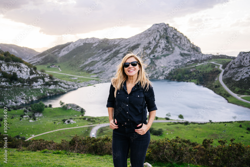 A beautiful caucasian blond woman in front of the camera wearing casual clothes in a mountainous landscape with lake