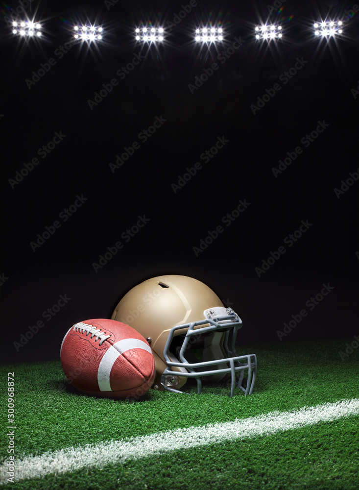 A gold football helmet and football on a grass field with stripe on dark background with stadium lights A gold football helmet and football on a grass field with stripe on dark background with stadium lights