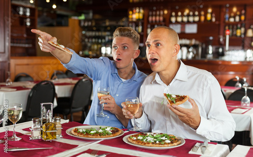 Two men watching sports while eating pizza in restaurant