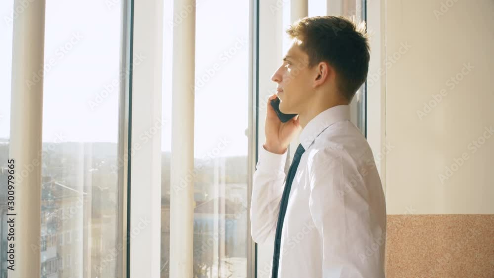 Young businessman in shirt and tie speaks by phone on in tall house on big windows background. Man work in big office. Slow motion. 4K. Young businessman in shirt and tie speaks by phone on in tall house on big windows background. Man work in big office. Slow motion. 4K.