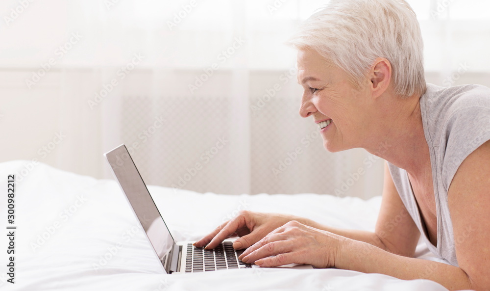 Cheerful senior lady typing on laptop computer, lying in bed Cheerful senior lady typing on laptop computer, lying in bed