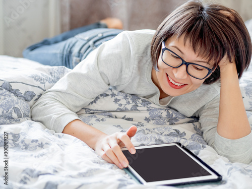 Woman in glasses are lying in bed with tablet. She is touching screen. Morning bedtime.