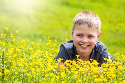 A boy with a smile is resting on a green lawn with yellow wildflowers in the sunshine
