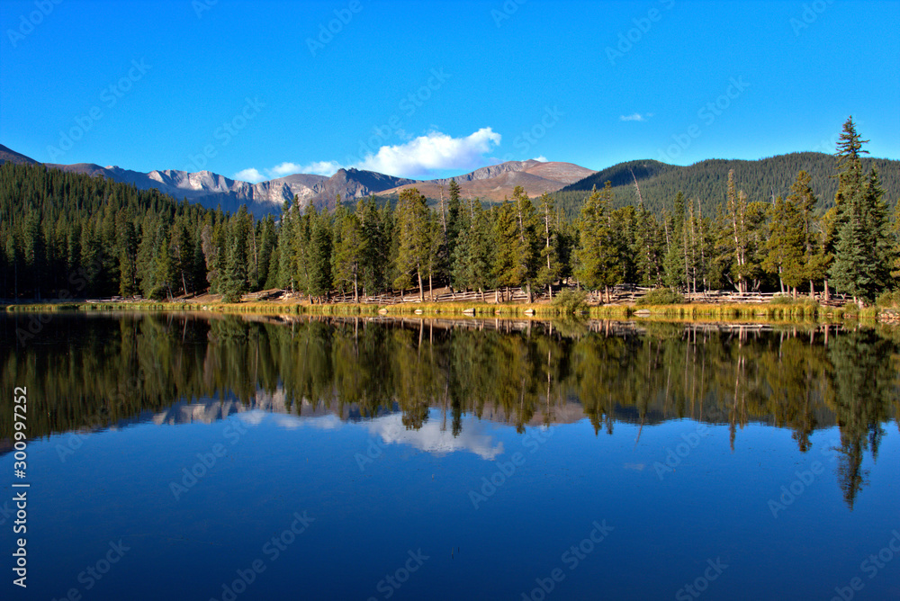 Breathtaking mountain lake with tall pine trees reflecting in the water with mountain peaks in the background with clear blue sky with puffy white clouds