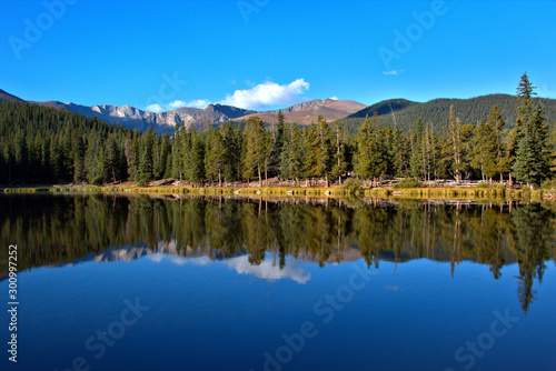 Breathtaking mountain lake with tall pine trees reflecting in the water with mountain peaks in the background with clear blue sky with puffy white clouds © ronm