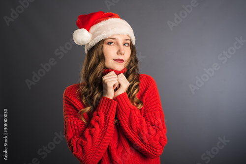 Cute and beautiful girl in red sweater and Santa hat on grey background. The concept of new year and Christmas  shopping. Place for your advertisement or text template
