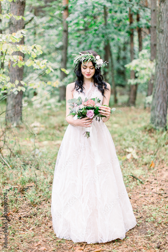 The bride with wreath on head in a stylish wedding dress in nature forest with a rustic bouquet. Photo shoot in pine forest in the style of fine art.