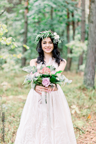 Portrait of a young beautiful bride with eucalypthus wreath on the head, with a rustic bouquet in the forest at wedding walk.