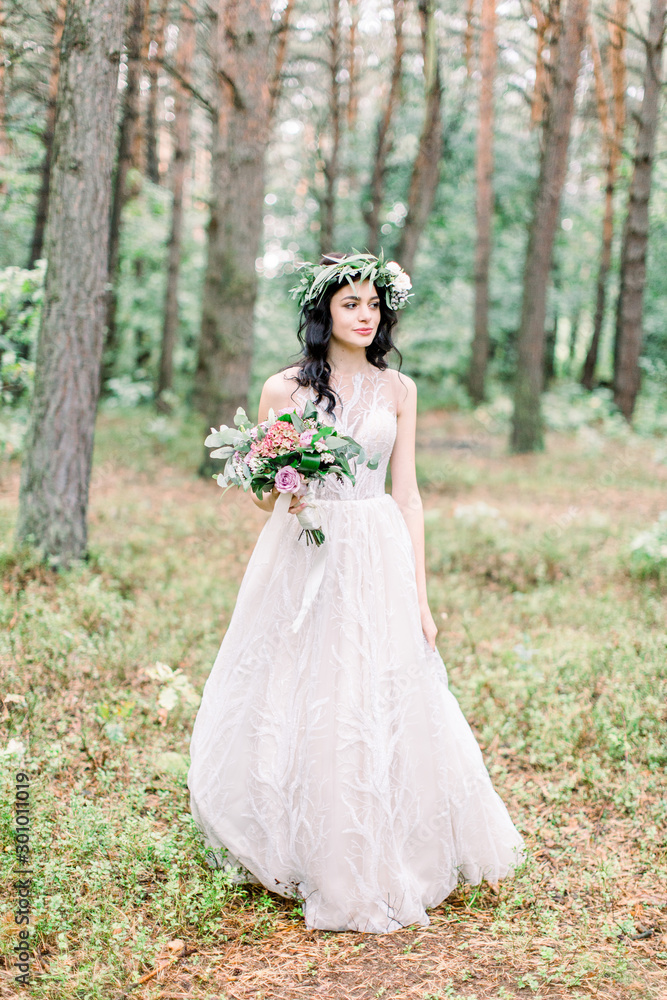 Full-length view of the charming bride in greenery wreath with the wedding bouquet looking aside in the spring forest.