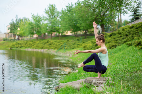 A young sports girl practices yoga on a green lawn by the river, yoga assans posture. Meditation and unity with nature © Kate