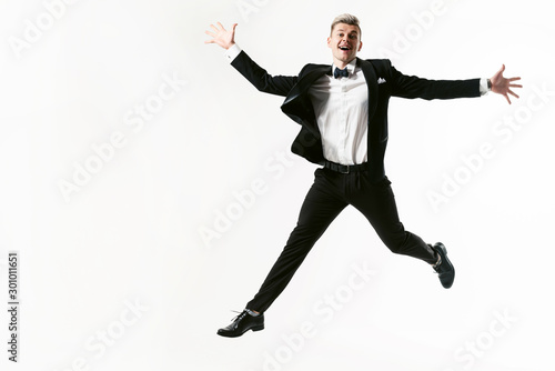 Portrait of young smiling handsome showman in tuxedo stylish black suit, studio shot jumping at white background. Businessman in jacket with bowtie