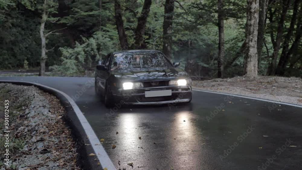 Black car starting drift with skidding and smoke on countryside road. Japanese car driving with slip and smoke on country highway on green forest trees background Black car starting drift with skidding and smoke on countryside road. Japanese car driving with slip and smoke on country highway on green forest trees background