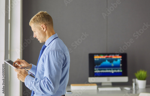 Portrait of smiling happy young entrepreneur standing at his office table and working on tablet computer