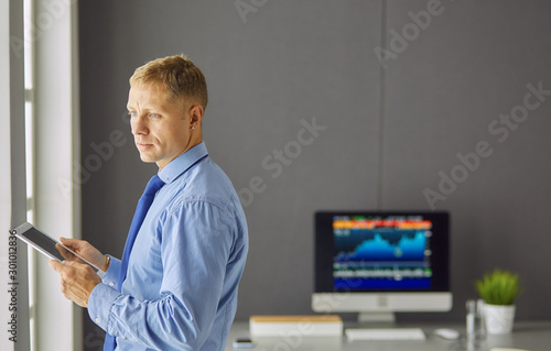 Portrait of smiling happy young entrepreneur standing at his office table and working on tablet computer